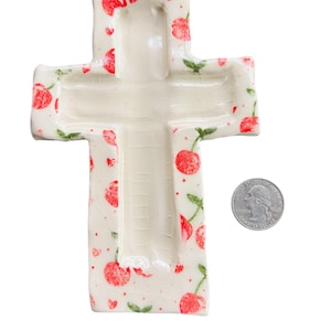 May include: A ceramic cross-shaped dish with a cream-colored base, adorned with red cherry patterns and green leaves. The dish has a glossy finish and a quarter coin is next to it for size comparison.