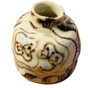 May include: A small, handcrafted ceramic vase with a rounded body and a narrow opening. The vase is off-white with brown and black decorative patterns and small blue accents. The rim is dark brown.