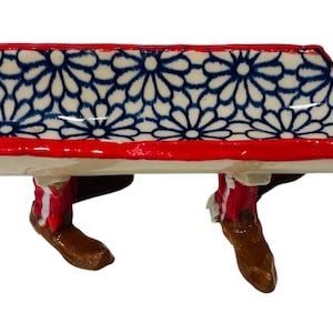 May include: A ceramic bench with a red trim and legs shaped like cowboy boots. The bench seat features a white background with a blue floral pattern. The boots are brown and red.