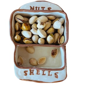 May include: A ceramic nut and shell dish filled with pistachios. The top section is labeled "NUTS" and holds shelled and unshelled pistachios. The bottom section, labeled "SHELLS", contains empty pistachio shells. The dish is off-white with brown trim.