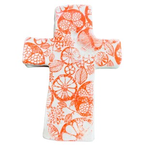 May include: A white cross-shaped decorative item with an orange floral pattern. The design includes stylized flowers, leaves, and fruit. The cross has rounded edges and a smooth surface, suitable for home decor or religious display.