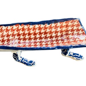 May include: A ceramic bench with a red and white houndstooth pattern seat and blue trim. The bench has blue and white patterned legs. The bench is a decorative item.