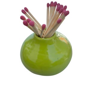 Puede incluir: Un pequeño jarrón redondo de cerámica verde lima lleno de cerillas de madera. Las cerillas tienen puntas rosas y están dispuestas para abanicarse desde el jarrón. El jarrón tiene un acabado brillante sobre un fondo blanco.