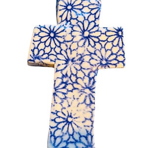 May include: A ceramic cross with a white base and a blue floral pattern. The cross is a light cream color with a repeating design of blue flowers. The cross is a religious symbol.