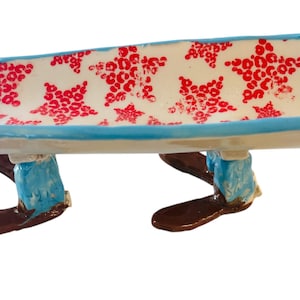 Puede incluir: Un banco de cerámica caprichoso con una superficie blanca estampada con formas de estrellas rojas. El banco tiene un borde azul y está sostenido por patas pintadas para parecerse a vaqueros azules y botas marrones.