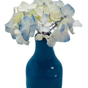 Könnte beinhalten: Ein Strauß weiß-blauer Hortensien in einer kleinen, dunkelblauen Vase. Die Blüten haben zarte Blütenblätter mit blauen Rändern und einem cremeweißen Zentrum. Die Vase hat eine einfache, abgerundete Form.