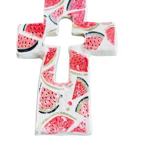 May include: A white cross-shaped decorative item with a watermelon pattern. The design features red watermelon slices with black seeds and green rind accents. The cross is a unique, summery decoration.