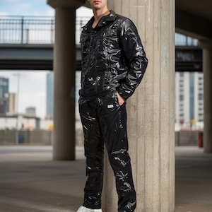 May include: A black, glossy two-piece suit, comprising a jacket and trousers. The jacket features a collar and pockets, while the trousers have a straight cut. The outfit is worn with white trainers. The setting is an urban environment.