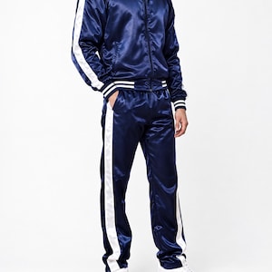 Könnte beinhalten: Ein marineblauer Trainingsanzug aus Satin, bestehend aus einer Reißverschlussjacke mit Stehkragen und einer passenden Hose. Beide Teile sind mit weißen Seitenstreifen sowie weiß gestreiften Rippbündchen an Ärmeln, Kragen und Saum versehen.