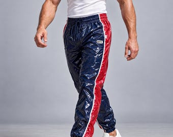 May include: Shiny navy blue track pants with a glossy finish, featuring a bold red vertical stripe down each side and a white drawstring waistband. A small rectangular patch with the text BMW is visible on the upper left thigh area.