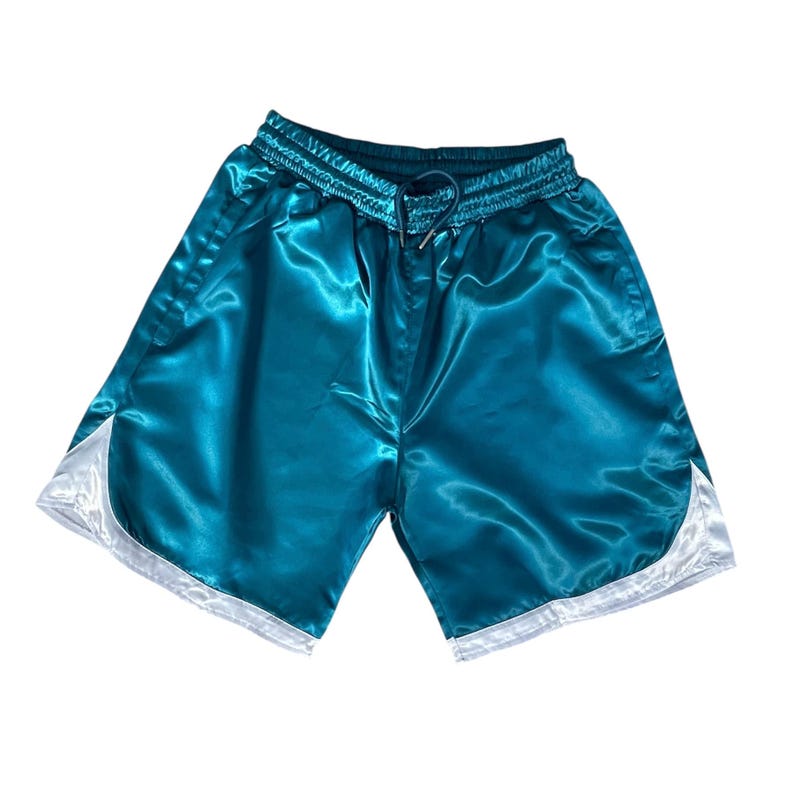 May include: Teal satin athletic shorts with a drawstring waist and white trim. These shorts have a shiny texture and a relaxed fit, suitable for sports or casual wear. The shorts are approximately 40 cm in length.