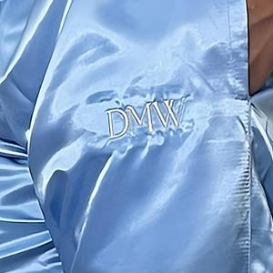 May include: Light blue satin pants with the embroidered letters "DMW". The pants have a shiny, smooth texture and a drawstring or elastic waistband. The fabric drapes in soft folds.