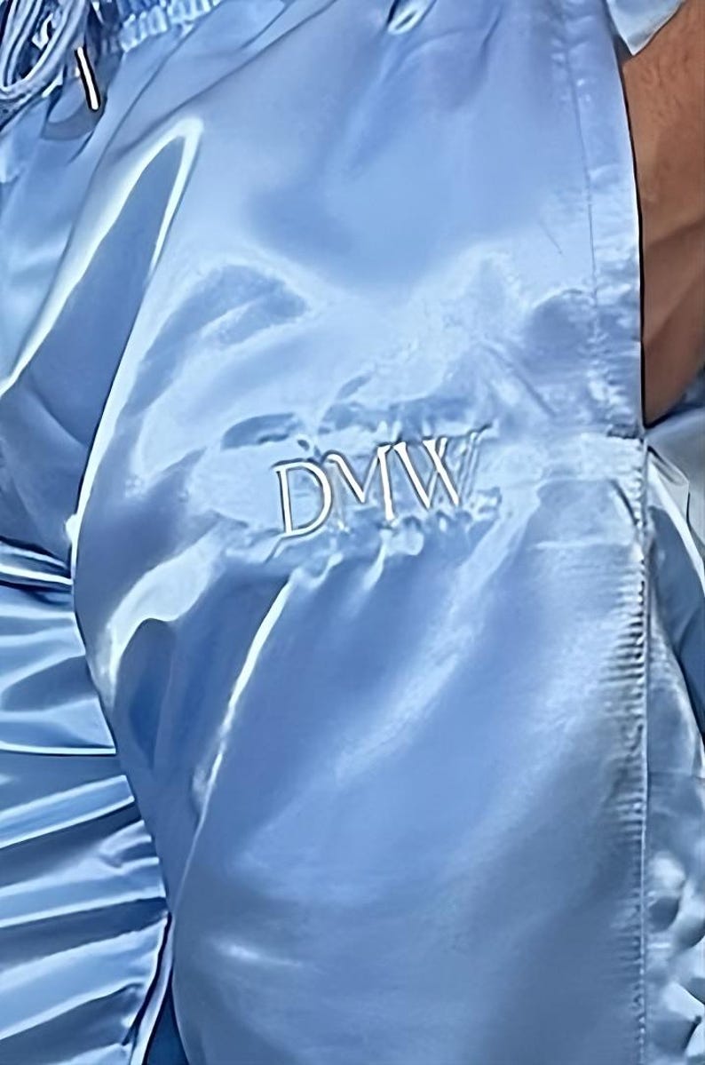 May include: Light blue satin pants with the embroidered letters "DMW". The pants have a shiny, smooth texture and a drawstring or elastic waistband. The fabric drapes in soft folds.