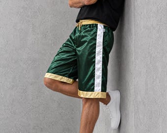 Könnte beinhalten: Sportshorts aus Satin in tiefem Smaragdgrün mit einem breiten, weißen vertikalen Seitenstreifen sowie einem goldfarbenen Bund und Saum. Die Shorts verfügen über einen goldenen Kordelzug in der Taille und werden mit einem schwarzen T-Shirt und weißen Sneakern kombiniert.