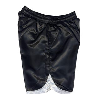 May include: Black satin shorts with a white trim along the bottom hem. The shorts have an elasticated waistband. The fabric has a shiny appearance. These athletic shorts are suitable for sports or casual wear.