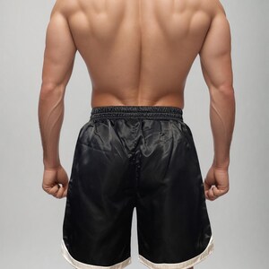 May include: Black satin boxing shorts with a white trim. The shorts are knee-length and have a drawstring waistband. The image shows the back of a person wearing the shorts, highlighting the athletic fit and design.