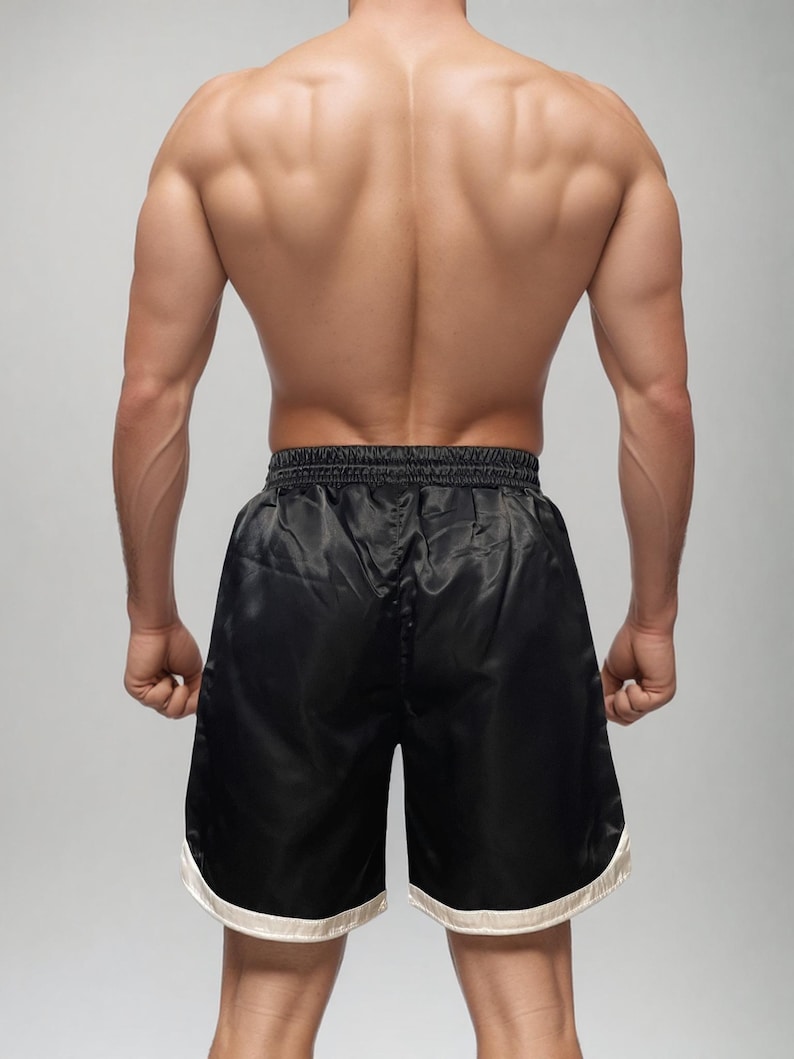 May include: Black satin boxing shorts with a white trim. The shorts are knee-length and have a drawstring waistband. The image shows the back of a person wearing the shorts, highlighting the athletic fit and design.