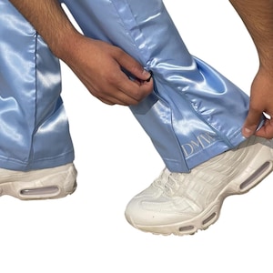 May include: Light blue satin pants with a zipper detail on the side. The pants have the embroidered letters "DMW" near the ankle. The person is wearing white sneakers.
