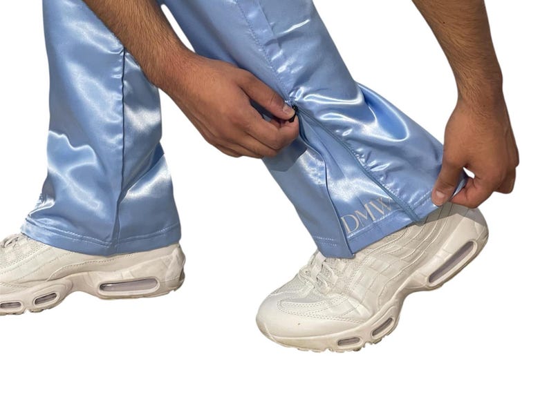 May include: Light blue satin pants with a zipper detail on the side. The pants have the embroidered letters "DMW" near the ankle. The person is wearing white sneakers.