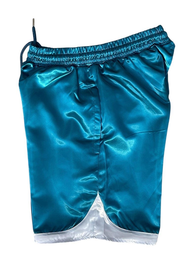 May include: Teal satin shorts with a drawstring waist and white trim along the bottom hem. The shorts have a shiny, reflective surface. The garment is designed for comfort and style.