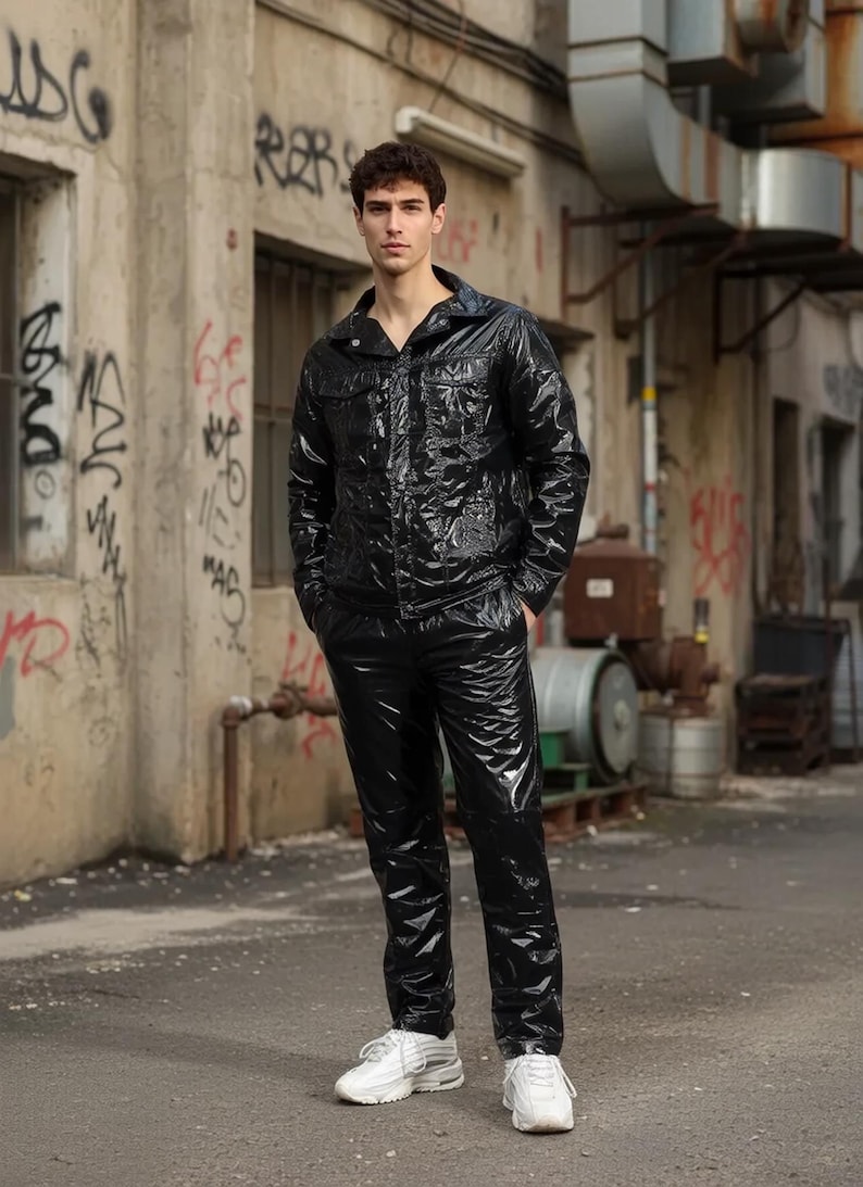 以下が含まれることがあります： 黒い光沢のあるフード付きジャケットと、お揃いのパンツを着た人。濡れたような外観のこの服装は、白いスニーカーと組み合わされています。背景には落書きのある建物があります。