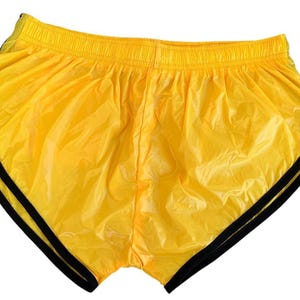 Op de afbeelding: Felgele gele sportbroek met zwarte bies. De short heeft een elastische tailleband en een glanzend, lichtgewicht materiaal. De short is ontworpen voor hardlopen of andere sportieve activiteiten.
