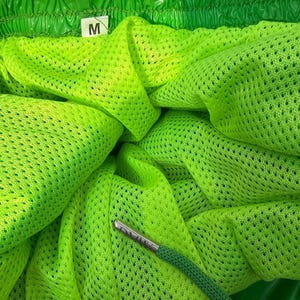 May include: Bright neon green athletic shorts featuring a fine mesh interior lining and an elasticized waistband. A white fabric tag with the letter M is attached to the inner seam. A green drawstring with a metallic silver aglet is visible.