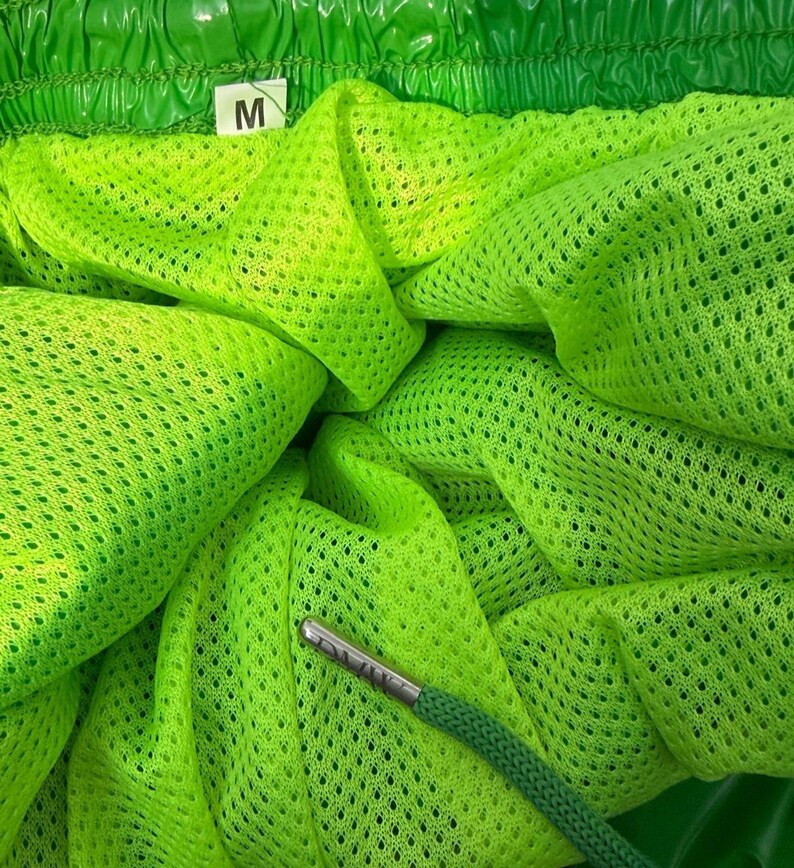 May include: Bright neon green athletic shorts featuring a fine mesh interior lining and an elasticized waistband. A white fabric tag with the letter M is attached to the inner seam. A green drawstring with a metallic silver aglet is visible.