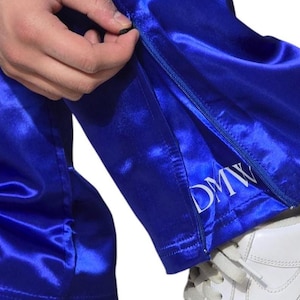 May include: Bright royal blue satin pants featuring a side zipper closure at the hem. The fabric has a reflective, smooth finish. The letters DMW are embroidered in white thread near the bottom edge of the leg opening above the zipper.
