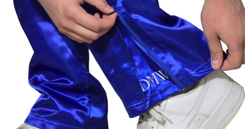 May include: Bright royal blue satin pants featuring a side zipper closure at the hem. The fabric has a reflective, smooth finish. The letters DMW are embroidered in white thread near the bottom edge of the leg opening above the zipper.