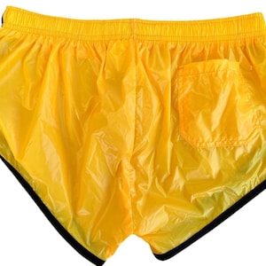 Op de afbeelding: Felge gele sportbroek met zwarte rand. De shorts hebben een klein zakje aan de achterkant en een elastische tailleband. Het materiaal lijkt glanzend, lichtgewicht nylon.