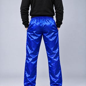 May include: Bright royal blue athletic pants with a shiny, satin-like finish and an elastic waistband. The pants feature a straight-leg cut and are paired with a black long-sleeved hooded sweatshirt and plain white sneakers.