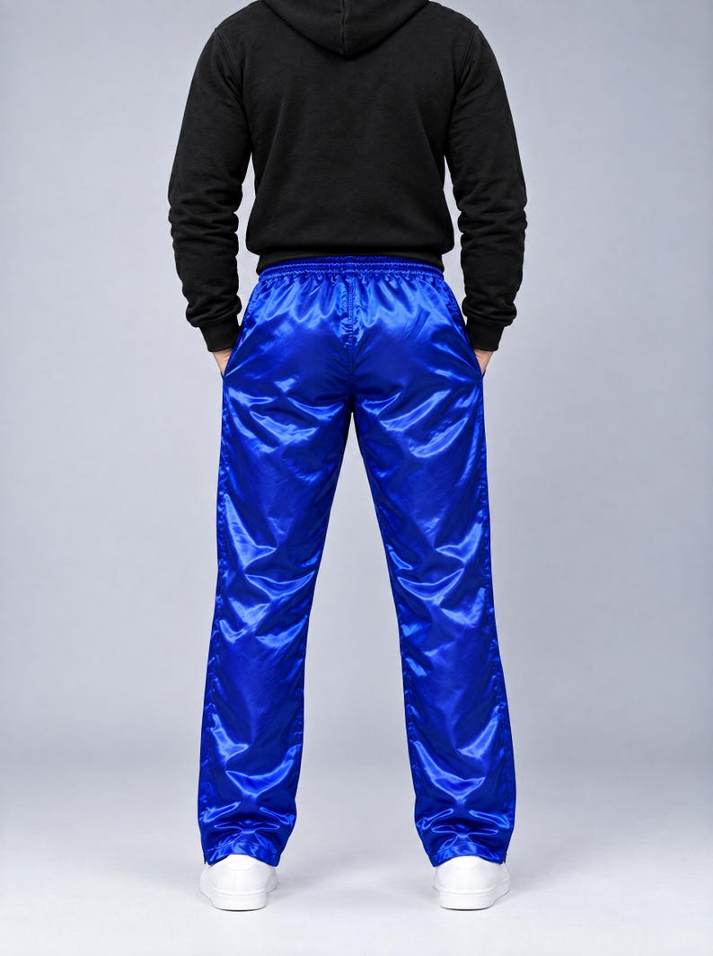 May include: Bright royal blue athletic pants with a shiny, satin-like finish and an elastic waistband. The pants feature a straight-leg cut and are paired with a black long-sleeved hooded sweatshirt and plain white sneakers.