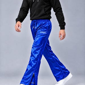 May include: Bright blue satin track pants featuring a drawstring waist, a white DMW logo on the upper thigh, and a matching DMW logo near the ankle. The pants have vertical side zippers at the bottom hem and are paired with a black hoodie and white sneakers.