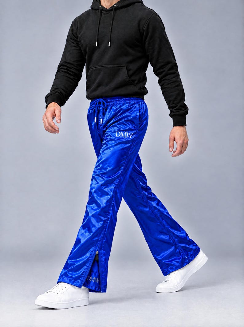 May include: Bright blue satin track pants featuring a drawstring waist, a white DMW logo on the upper thigh, and a matching DMW logo near the ankle. The pants have vertical side zippers at the bottom hem and are paired with a black hoodie and white sneakers.