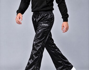May include: Black satin track pants featuring a drawstring waistband, side zippers along the legs, and the white text DMW printed on the upper thigh and near the bottom hem. The pants have a shiny finish and a straight-leg silhouette.