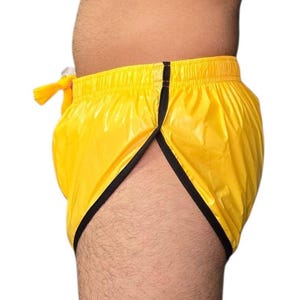 Op de afbeelding: Gele sportbroek met zwarte rand. De shorts hebben een trekkoord en een splitzoom. Het materiaal lijkt een glanzende, lichtgewicht stof te zijn. De shorts worden van opzij getoond.