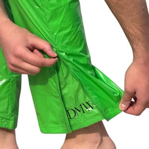 May include: Bright lime green pants made of a shiny, water-resistant material. The lower leg features a vertical zipper opening along the side seam. Near the bottom hem, the letters DMW are printed in a bold, black, serif font on the fabric.