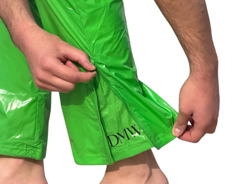 May include: Bright lime green pants made of a shiny, water-resistant material. The lower leg features a vertical zipper opening along the side seam. Near the bottom hem, the letters DMW are printed in a bold, black, serif font on the fabric.