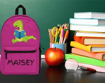 May include: A pink backpack featuring a green dinosaur reading a book. The backpack has a black zipper and the name "MAISEY" is printed on the front.