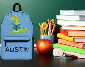 May include: A light blue backpack featuring a green dinosaur wearing glasses and reading a book. The backpack has a black zipper and the name "AUSTIN" is written in black on the front.