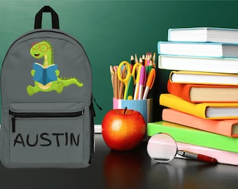 May include: Grey backpack featuring a green dinosaur wearing spectacles and reading a blue book. The backpack has a black zip and the name "AUSTIN" is written in black on the front.