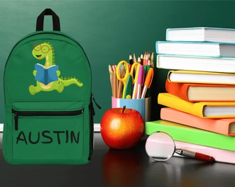 May include: Green canvas backpack featuring a cartoon dinosaur wearing glasses and reading a book. The backpack has a black zipper and a black name tag that reads "AUSTIN".