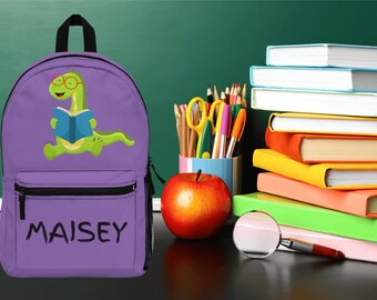 May include: Purple backpack featuring a green dinosaur reading a book. The backpack has a black zipper and the name "MAISEY" is written in black on the front.