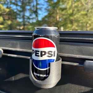 Puede incluir: Una lata de Pepsi Zero Sugar negra y plateada en un portavasos gris. La lata tiene el logotipo de Pepsi en rojo, blanco y azul. Las palabras "ZERO SUGAR" están impresas en la lata.
