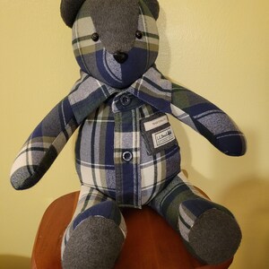 May include: A stuffed bear crafted from blue, green, and white plaid flannel fabric. The bear features dark gray felt ears and foot pads, a black nose, and a chest pocket with a label reading L.L.Bean and Traditional Fit. It has a buttoned collar.