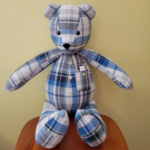 May include: A handmade teddy bear with a blue, white, and black plaid pattern. The bear has button eyes, a black nose, and a small pocket on its chest. It is sitting on a wooden surface.