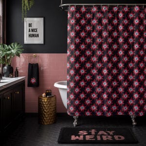 May include: A black shower curtain with a repeating red and blue floral pattern. A matching black hand towel with the same floral design hangs on a gold towel rack. A black bath mat with the words "STAY WEIRD" is on the floor.