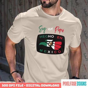 Könnte beinhalten: Ein beiges kurzärmeliges T-Shirt mit dem Text Soy un Papá in grüner und roter Schreibschrift. Darunter zeigt eine schwarze rechteckige Grafik den Text HECHO EN MEXICO mit einem Adler-Symbol in den Farben Grün, Weiß und Rot.