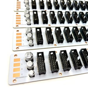Op de afbeelding: Vier witte rechthoekige printplaten parallel gerangschikt. Elke printplaat is voorzien van drie cilindrische zilverkleurige condensatoren, een reeks zwarte meerpolige headers en koperen contactpunten met spanningsmarkeringen en serienummers.